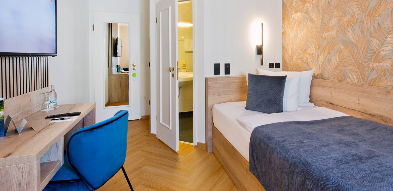 Einzelzimmer: rechts ist ein Bett; daneben ist eine offene Tür, die zum Bad führt; links befindet sich ein Schreibtisch mit blauen Schreibtischstuhl und darüber einem Fernseher