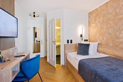 Einzelzimmer: rechts ist ein Bett; daneben ist eine offene Tür, die zum Bad führt; links befindet sich ein Schreibtisch mit blauen Schreibtischstuhl und darüber einem Fernseher