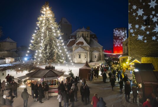 Veranstaltung Weihnachtszauber auf der Burg Querfurt, Marktstände und Menschen im Schnee, ein Weihnachtsbaum mit Lichterkette und auf die Burgmauern sind übergroße Schneeflocken projiziert