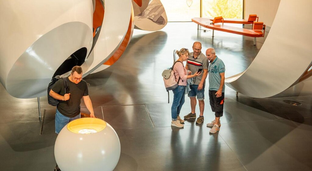 Sichtbar ist der Raum der Dauerausstellung der Arche Nebra mit verschiedenen Elementen. Drei Personen stehen in der Mitte des Raums und schauen gemeinsam auf ein Handy. Eine weitere Person schaut auf Ausstellungsstück.