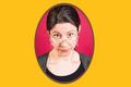 Eine Frau blickt skeptisch über den Rand ihrer Brille nach vorn. Der Hintergrund ist gelb.