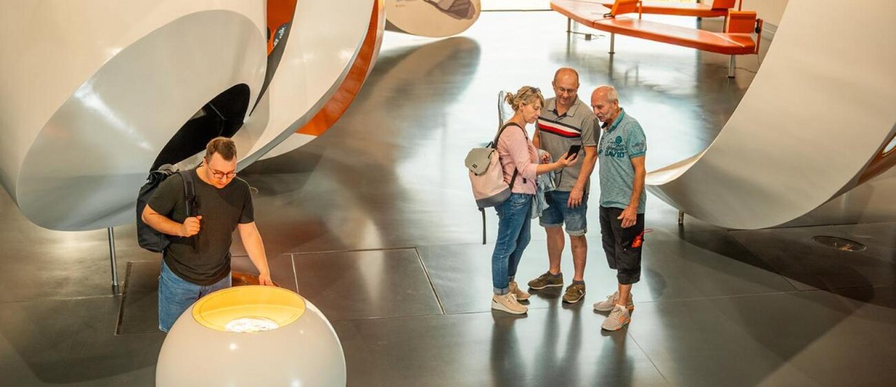 Sichtbar ist der Raum der Dauerausstellung der Arche Nebra mit verschiedenen Elementen. Drei Personen stehen in der Mitte des Raums und schauen gemeinsam auf ein Handy. Eine weitere Person schaut auf Ausstellungsstück.