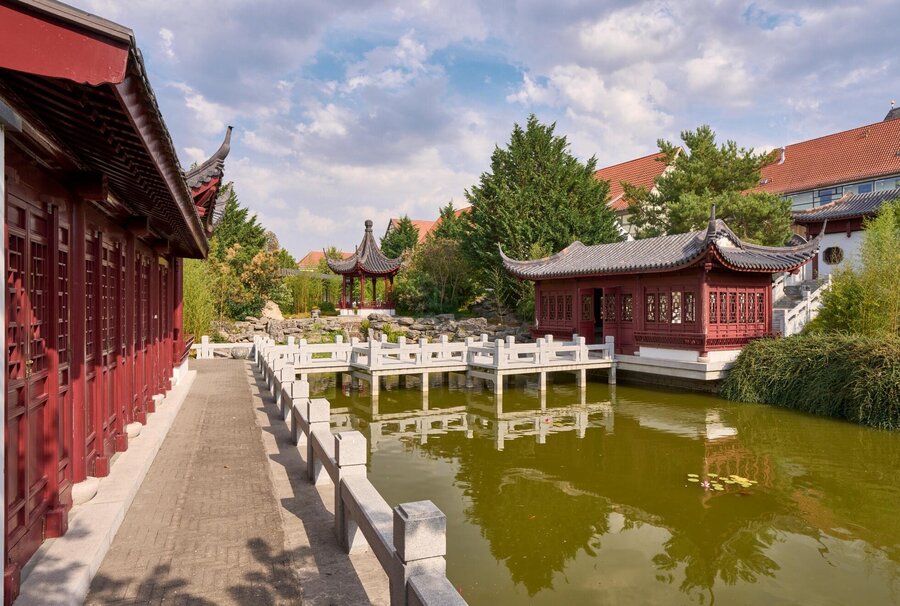 Chinesischer Garten mit roten Häusern im chinesischen Stil mit weißer Zick-Zack-Brücke über einem Teich