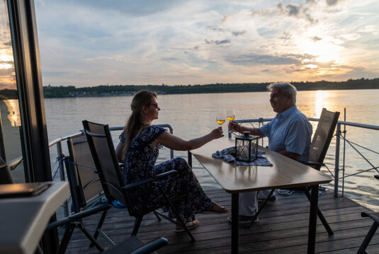 Ein Paar stößt mit Wein auf der Terrasse eines Hausbootes auf dem Geiseltalsee an, im Hintergrund geht die Sonne unter.