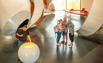Sichtbar ist der Raum der Dauerausstellung der Arche Nebra mit verschiedenen Elementen. Drei Personen stehen in der Mitte des Raums und schauen gemeinsam auf ein Handy. Eine weitere Person schaut auf Ausstellungsstück.